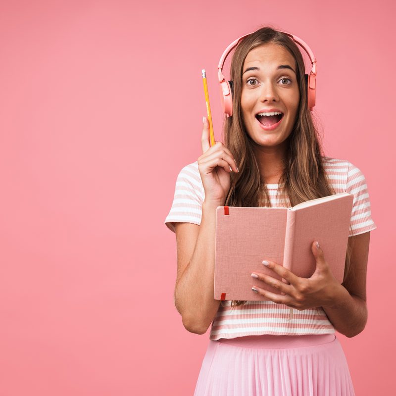 image-of-excited-beautiful-girl-writing-down-notes-at-diary-book-and-listening-to-music-with-headphones-isolated-over-pink-wall.jpg